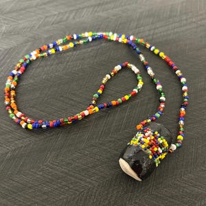 Puede incluir: Un collar hecho a mano con un colgante cilíndrico y una cadena de cuentas. El colgante es negro con cuentas coloridas. La cadena del collar está hecha de pequeñas cuentas multicolores en rojo, amarillo, azul y blanco. El collar se muestra sobre una superficie oscura.