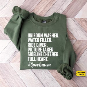 May include: A green sweatshirt with white text that reads "UNIFORM WASHER. WATER FILLER. RIDE GIVER. PICTURE TAKER. SIDELINE CHEERER. FULL HEART. #Sportsmom".