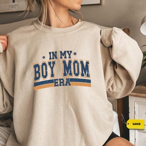 May include: A beige sweatshirt with the text "IN MY BOY MOM ERA" in navy blue and orange. The text is in a bold, blocky font with a star on each side of the phrase. The sweatshirt has long sleeves and a crew neck.