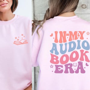 a woman wearing a pink sweatshirt with the words in my audio book era printed on