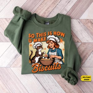 May include: Olive green sweatshirt with a graphic that reads "SO THIS IS HOW U MAKE Biscuits." The graphic features a woman and a cat wearing chef hats, with the woman mixing ingredients in a bowl. The sweatshirt has long sleeves and a crew neck.