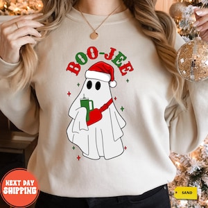 May include: Cream-colored sweatshirt featuring a cartoon ghost wearing a Santa hat and holding a green drink. The text "BOO-JEE" is above the ghost in red and green. Red and green stars are scattered around the ghost.