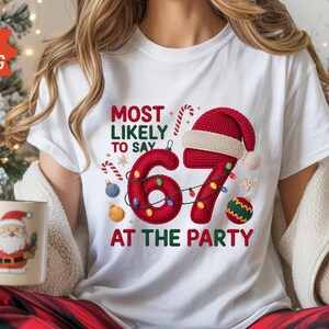 May include: White t-shirt with a festive Christmas design. The shirt displays the text "MOST LIKELY TO SAY 67 AT THE PARTY" in red, accompanied by a Santa hat, candy canes, and baubles. A mug with a Santa Claus image is also visible.