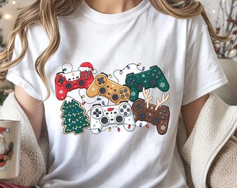 Könnte beinhalten: Weißes T-Shirt mit einem festlichen Design aus bunten Gamecontrollern, die mit festlichen Elementen dekoriert sind. Die Controller sind rot, gold, grün, braun und weiß, mit einem Weihnachtsbaum und Lichterketten. Auf einem Controller ist ein Weihnachtsmannhut.