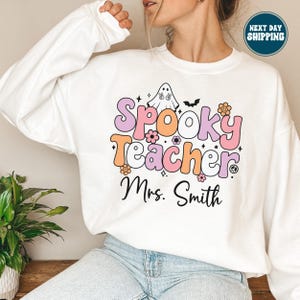 May include: White sweatshirt with the text "Spooky Teacher Mrs. Smith" in a retro font. The design includes a ghost, flowers, and a bat. The sweatshirt is a crew neck style.