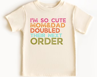 May include: Cream-colored t-shirt with the text "I'M SO CUTE MOM&DAD DOUBLED THEIR NEXT ORDER" in pink, orange, teal, and green. The shirt is made of soft material and has short sleeves.