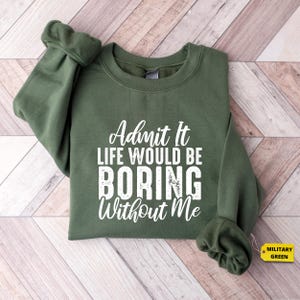 May include: A military green sweatshirt featuring the white text "Admit It LIFE WOULD BE BORING Without Me". The sweatshirt has a crew neck and long sleeves. The tag reads "MILITARY GREEN".