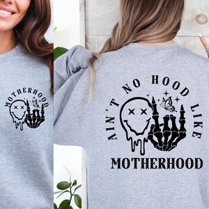 May include: Gray sweatshirt with a black graphic print on the front and back. The front graphic is a skull with a smiley face and a butterfly. The back graphic says "Ain't no hood like motherhood" with a skull with a smiley face and a butterfly.