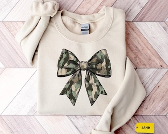 May include: A beige sweatshirt with a large camouflage bow printed on the front. The sweatshirt has a tag that says "SAND" on it.