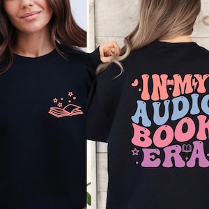 May include: Black sweatshirt with a small coral-colored book and star design on the front. The back of the sweatshirt features the text "IN MY AUDIO BOOK ERA" in retro-style lettering with pink, blue, and purple colors.