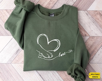 May include: A green sweatshirt with a white outline of a hand holding a paw inside a heart with the word "love" written below. The sweatshirt is folded and laid on a wooden surface.