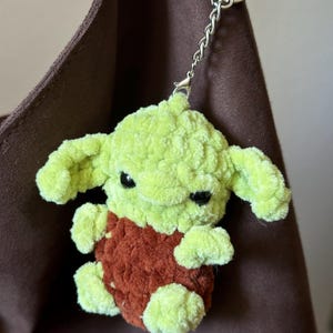 Pu&ograve; includere: Un portachiavi all'uncinetto fatto a mano, verde e ruggine, raffigurante una piccola creatura con grandi orecchie. Il portachiavi ha una catena e un anello argentati per attaccarlo alle chiavi o a una borsa. Il peluche &egrave; morbido e dettagliato.