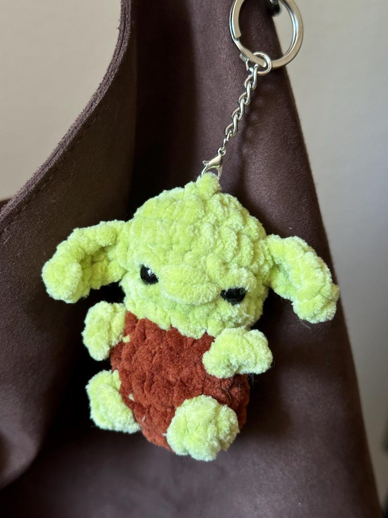 Pu&ograve; includere: Un portachiavi all'uncinetto fatto a mano, verde e ruggine, raffigurante una piccola creatura con grandi orecchie. Il portachiavi ha una catena e un anello argentati per attaccarlo alle chiavi o a una borsa. Il peluche &egrave; morbido e dettagliato.