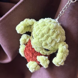 Pu&ograve; includere: Un portachiavi fatto a mano, all'uncinetto in verde chiaro e arancione, con una piccola creatura di peluche. Il portachiavi ha una catena e un anello argentati. La creatura ha un corpo verde chiaro e un gilet arancione.