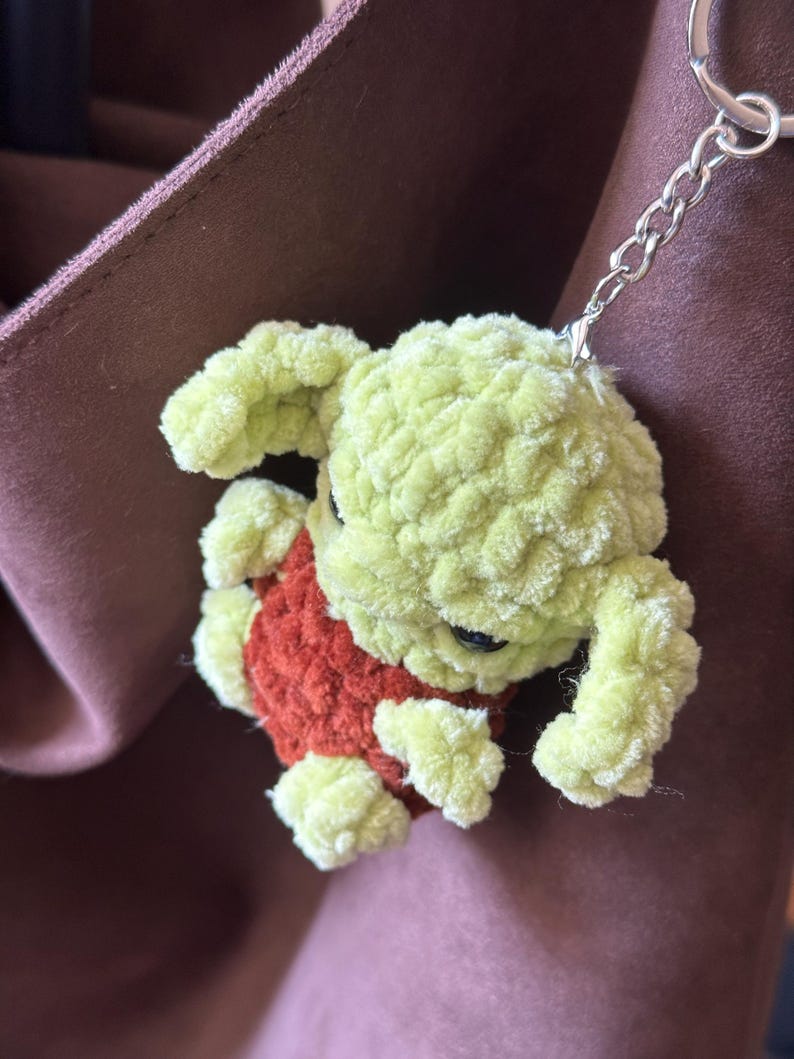Pu&ograve; includere: Un portachiavi fatto a mano, all'uncinetto in verde chiaro e arancione, con una piccola creatura di peluche. Il portachiavi ha una catena e un anello argentati. La creatura ha un corpo verde chiaro e un gilet arancione.