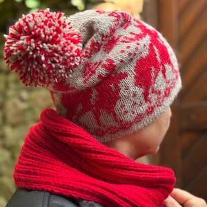 Könnte beinhalten: Eine gestrickte Wintermütze mit einem roten und grauen Rentier-Muster und einem großen rot-weißen Bommel. Ein passender roter Strickschal ist um den Hals gewickelt. Mütze und Schal sind aus weichem, warmem Material.
