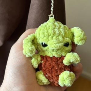 Pu&ograve; includere: Un portachiavi all'uncinetto fatto a mano, verde e marrone, raffigurante un personaggio con grandi orecchie e occhi neri. Il portachiavi &egrave; attaccato a un anello di metallo argentato. Il peluche &egrave; realizzato in filato morbido e strutturato.