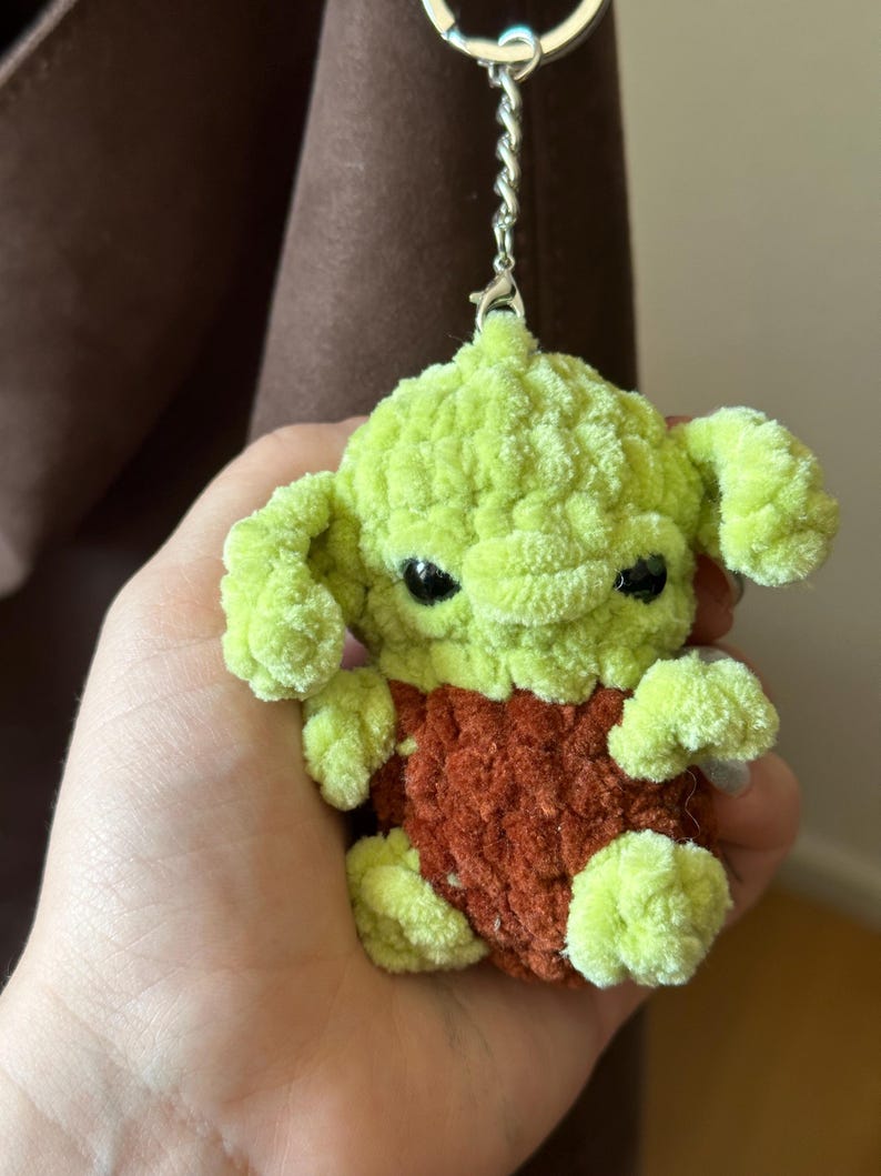 Pu&ograve; includere: Un portachiavi all'uncinetto fatto a mano, verde e marrone, raffigurante un personaggio con grandi orecchie e occhi neri. Il portachiavi &egrave; attaccato a un anello di metallo argentato. Il peluche &egrave; realizzato in filato morbido e strutturato.