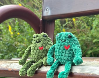 Op de afbeelding: Twee handgemaakte gehaakte kikkerknuffels. Eén is donkergroen, de andere heldergroen. Beide hebben zwarte ogen en een klein rood hart. De kikkers zitten op een houten bank met een vage groene achtergrond.