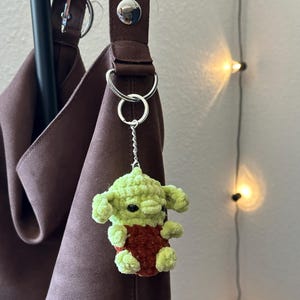 Pu&ograve; includere: Un portachiavi a forma di personaggio verde e rosso all'uncinetto &egrave; appeso a un cinturino di una borsa marrone. Il portachiavi presenta una piccola figura di peluche con grandi orecchie e occhi neri. Il cinturino della borsa &egrave; di colore marrone scuro e ha finiture argentate.