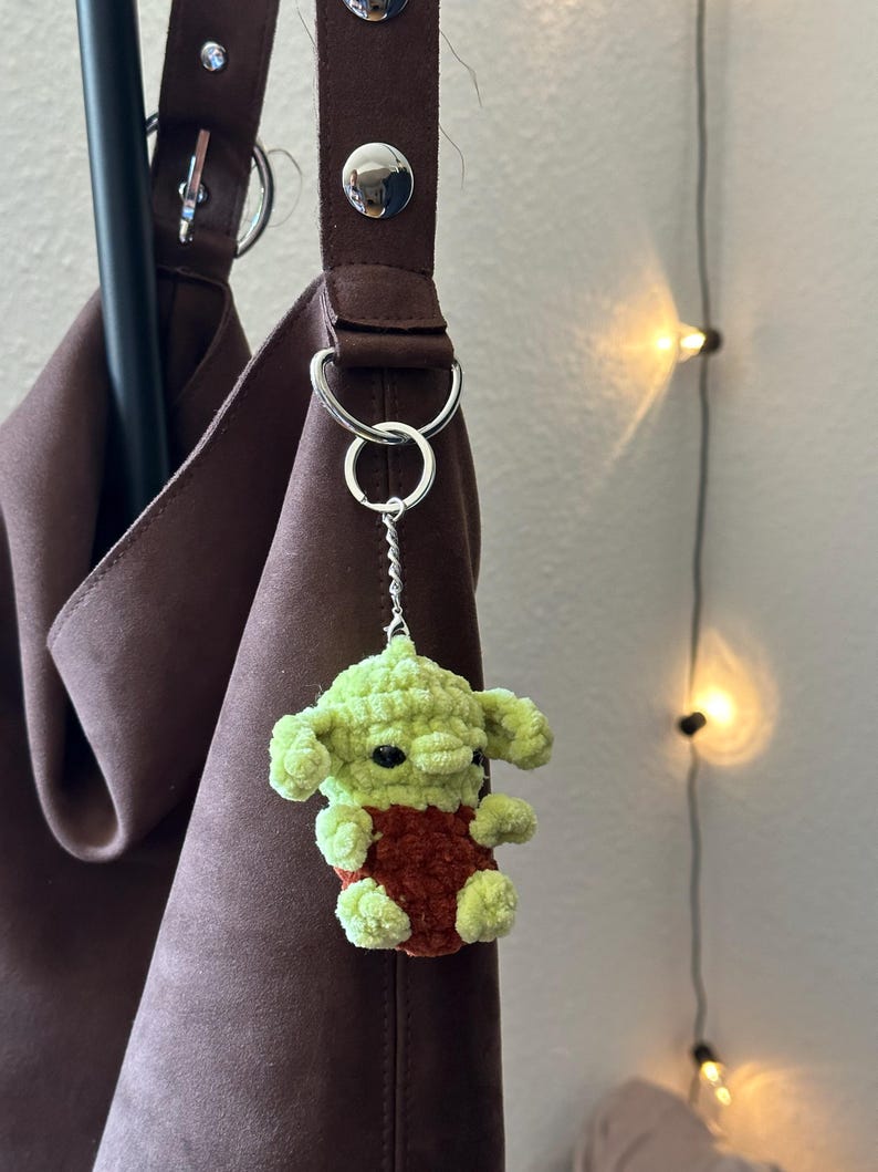 Pu&ograve; includere: Un portachiavi a forma di personaggio verde e rosso all'uncinetto &egrave; appeso a un cinturino di una borsa marrone. Il portachiavi presenta una piccola figura di peluche con grandi orecchie e occhi neri. Il cinturino della borsa &egrave; di colore marrone scuro e ha finiture argentate.