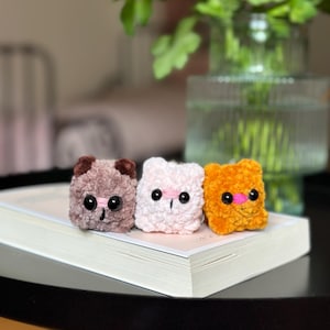 Puede incluir: Tres pequeñas figuras de animales de ganchillo hechas a mano. Las figuras son un oso marrón claro, un gato rosa y un oso naranja. Cada uno tiene ojos negros y una nariz rosa. Están sobre un libro blanco.