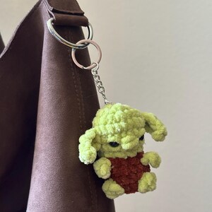 Pu&ograve; includere: Un portachiavi all'uncinetto verde e marrone raffigurante un piccolo personaggio con grandi orecchie e occhi scuri. Il portachiavi &egrave; attaccato a una catena e un anello argentati, appeso a un cinturino marrone.