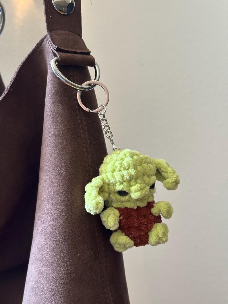 Pu&ograve; includere: Un portachiavi all'uncinetto verde e marrone raffigurante un piccolo personaggio con grandi orecchie e occhi scuri. Il portachiavi &egrave; attaccato a una catena e un anello argentati, appeso a un cinturino marrone.