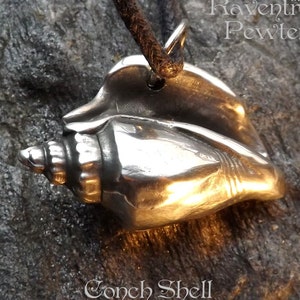 May include: A silver conch shell pendant, with a detailed, realistic design. The pendant is hanging from a brown leather cord. The text "Raventree Pewter" is visible in the background.