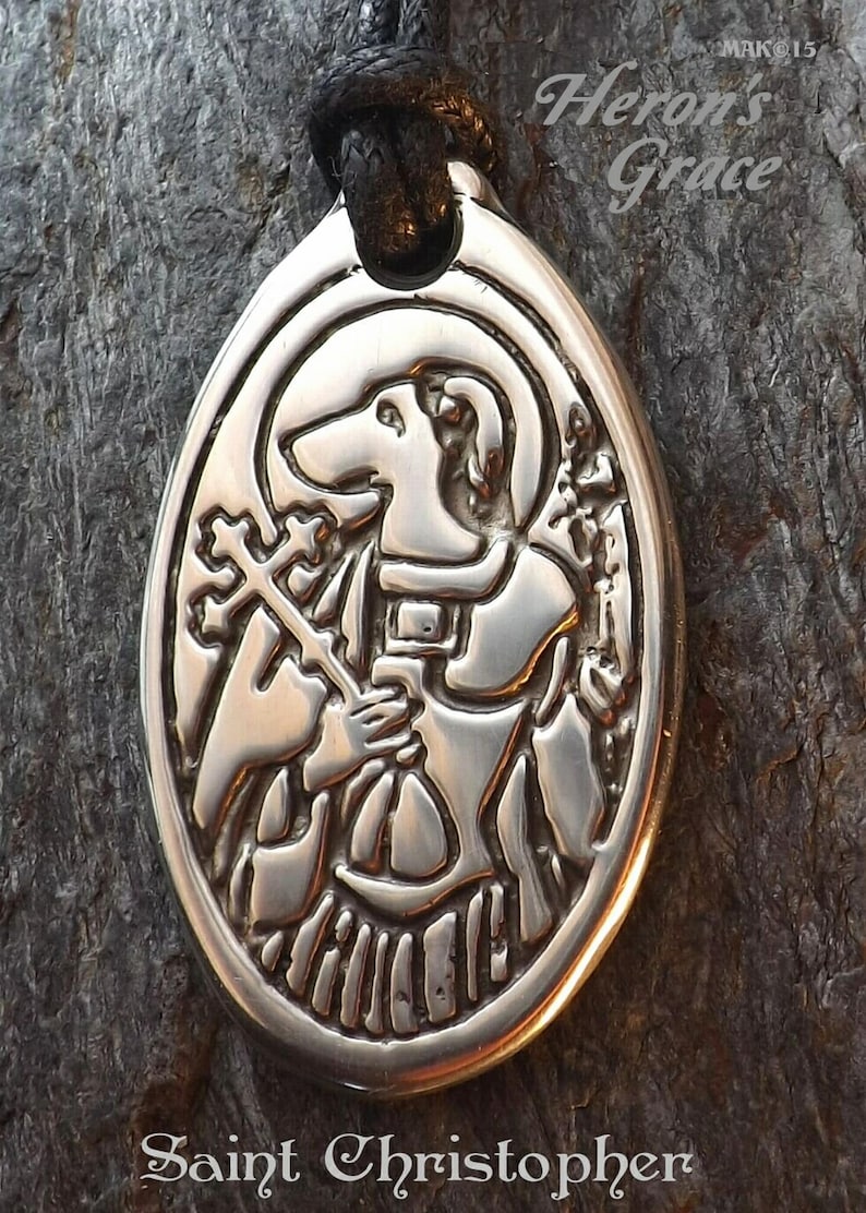 May include: A silver oval pendant with a detailed image of Saint Christopher carrying a staff and a child. The pendant is on a black cord and has the text "Heron's Grace" and "Saint Christopher" on it.