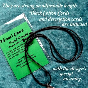 May include: A black cotton cord necklace with a pewter pendant that says "Heron's Grace Hand Poured". The pendant is attached to a green card with the text "Pewter with a message I design, mold, and hand pour each piece with the finest lead free pewter. They are to inspire and encourage lives with unique messages. Mark Kroh - Master Wire Smith - Made by hand in America Heron'sGrace.com". The card is in a clear plastic bag. The text "They are strung on adjustable length Black Cotton Cords and description cards are included with the design's special meaning." is written above the image.