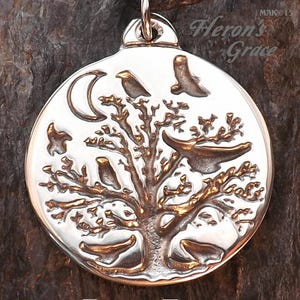 May include: A silver pendant featuring a detailed tree with birds and a crescent moon. The pendant is round and has a textured surface. The text "Raven Tree" is printed below the pendant.