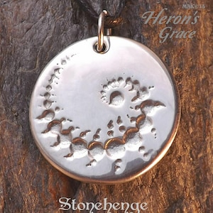 May include: Silver pendant necklace with a circular design featuring a raised, textured image of Stonehenge. The pendant is on a black cord and has the text "Heron's Grace" above it and "Stonehenge" below it.