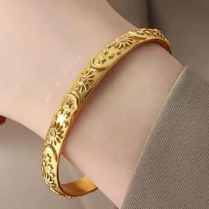 May include: A gold-toned bangle bracelet featuring a repeating embossed pattern of suns, crescent moons, and small stars. The surface of the bracelet is polished and includes small, clear, circular accents embedded within the center of each sun motif.
