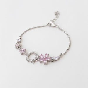 Puede incluir: Una pulsera de cadena en tono plateado con una serie de dijes que incluyen una flor central de cristal rosa, un contorno con forma de gato y detalles florales adicionales con pequeños cristales transparentes y piedras rosas, con cierre de mosquetón.