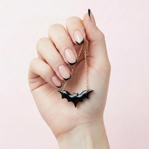 Puede incluir: Un collar con colgante de murciélago negro con una cadena dorada. El colgante de murciélago es negro brillante y tiene un diseño de alas detallado. El collar se sostiene sobre un fondo rosa claro.