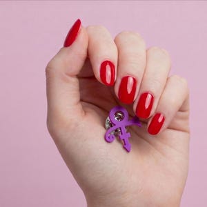 Puede incluir: Un pin de símbolo morado, sostenido en una mano con esmalte de uñas rojo, sobre un fondo rosa. El pin es un glifo estilizado, probablemente un logotipo o emblema. El pin es de metal.
