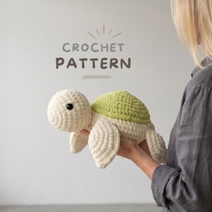 May include: A crocheted turtle toy with a cream-colored body and a green shell. The turtle has black eyes and is being held by a person. The text "CROCHET PATTERN" is above the turtle.