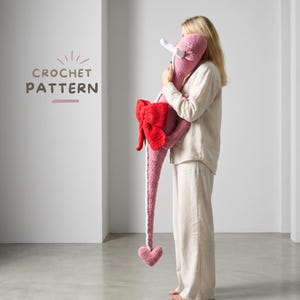 Peut inclure: Un dragon rose au crochet avec un nœud rouge et une queue en forme de cœur. Le dragon a des cornes blanches et une longue queue. Le texte "CROCHET PATTERN" est visible en haut à gauche.
