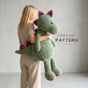 Könnte beinhalten: Ein großer, handgefertigter Häkeldrachen in Grün- und Rosatönen. Das Plüschtier hat einen langen Schwanz, vier Beine und einen Kopf mit kleinen Hörnern. Der Text "CROCHET PATTERN" ist im Bild sichtbar.