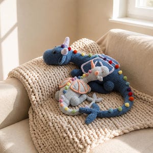 May include: Two crocheted dragon plush toys in blue and gray with rainbow accents rest on a beige blanket. The larger dragon has a long tail and colorful pom-poms. The smaller dragon has a horn and a rainbow-colored spine.
