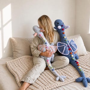May include: Two handmade crochet dragon plushies. One is light blue with pink wings, the other is blue with white wings. Both have rainbow-colored pom-poms down their spines and are on a bed with a beige blanket.