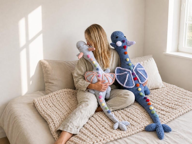 May include: Two handmade crochet dragon plushies. One is light blue with pink wings, the other is blue with white wings. Both have rainbow-colored pom-poms down their spines and are on a bed with a beige blanket.