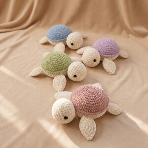 May include: A collection of handmade crochet turtle toys in various pastel colors. The turtles have cream-colored bodies, with shells in blue, green, purple, and pink. The toys are arranged on a beige fabric background.