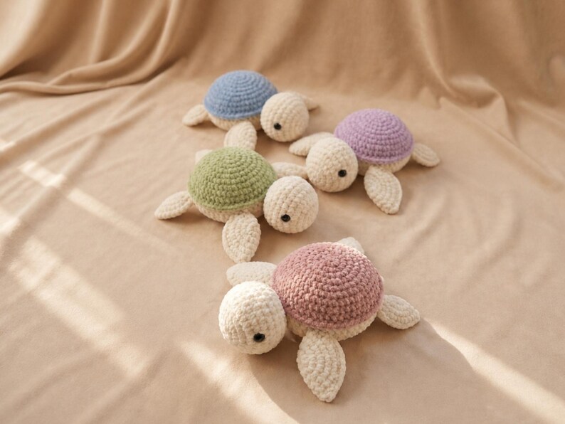 May include: A collection of handmade crochet turtle toys in various pastel colors. The turtles have cream-colored bodies, with shells in blue, green, purple, and pink. The toys are arranged on a beige fabric background.