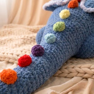 May include: A blue crocheted dinosaur-shaped pillow with colorful pom-pom accents. The plush toy features a long tail and a textured, handmade appearance. The pom-poms are in shades of orange, red, purple, blue, green, and yellow.
