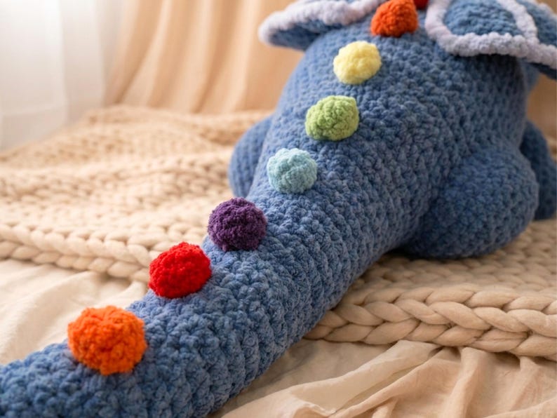 May include: A blue crocheted dinosaur-shaped pillow with colorful pom-pom accents. The plush toy features a long tail and a textured, handmade appearance. The pom-poms are in shades of orange, red, purple, blue, green, and yellow.