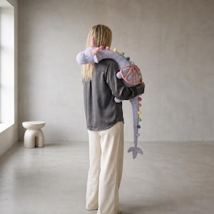 May include: A person holding a large, gray stuffed animal shaped like a dragon. The dragon has colorful pom-poms along its spine and tail, with pink and purple accents. The person is wearing a dark gray shirt and cream-colored pants.