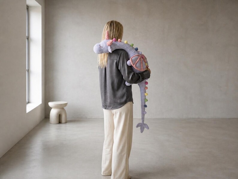 May include: A person holding a large, gray stuffed animal shaped like a dragon. The dragon has colorful pom-poms along its spine and tail, with pink and purple accents. The person is wearing a dark gray shirt and cream-colored pants.