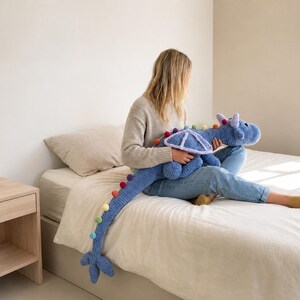 May include: A large, blue crocheted dragon plush toy with rainbow pom-pom accents along its tail and back. The dragon has a detailed head and wings. The toy is soft and cuddly, perfect for children or as a decorative item.